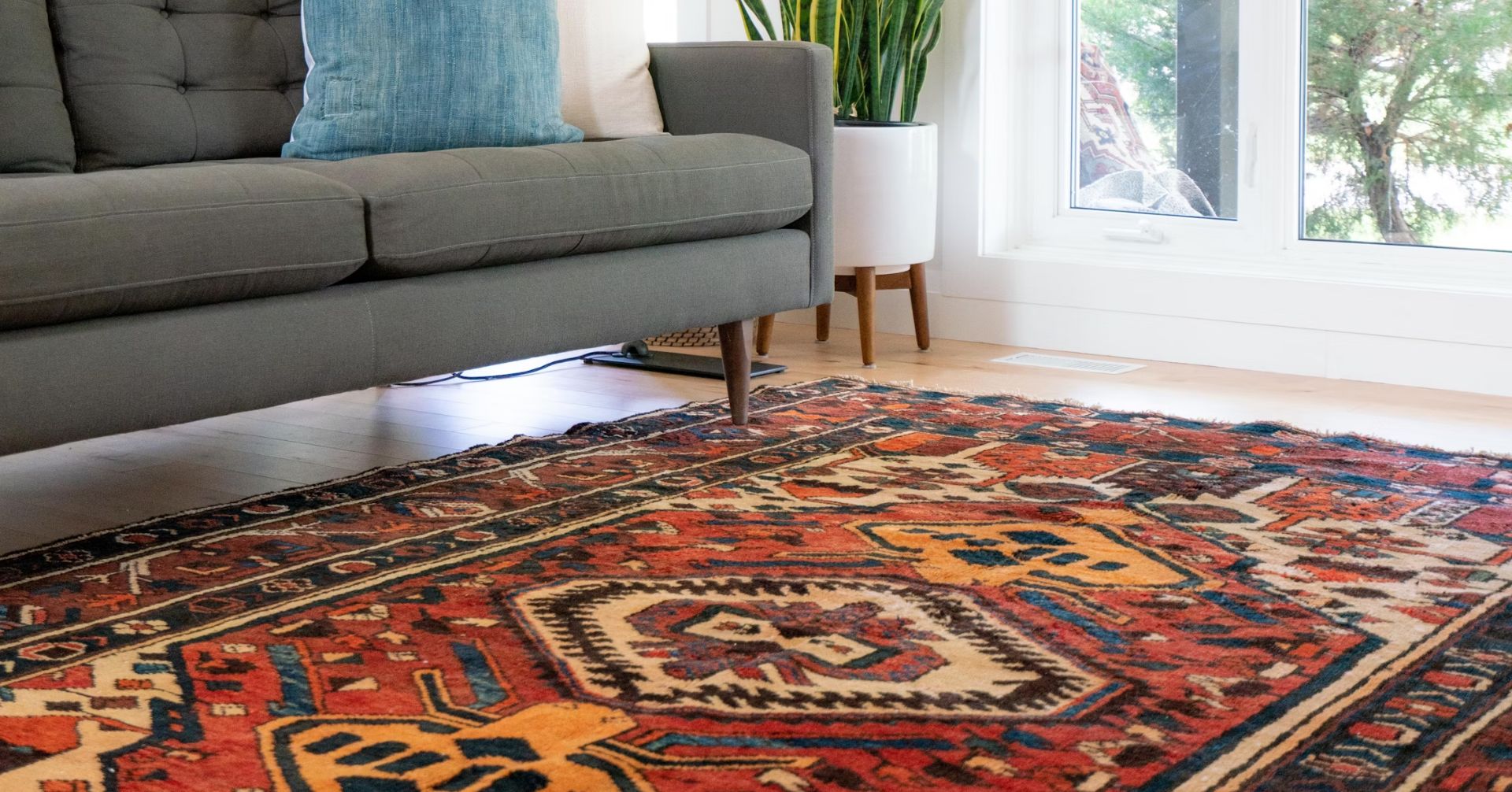 A bright and colourful oriental rug in a living room under a grey sofa with a blue cushion with a potted plant next to the window